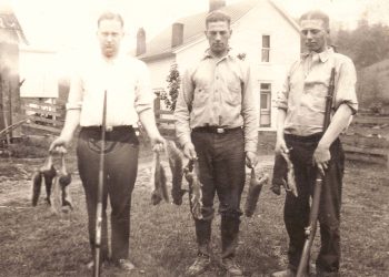 A vintage black and white photo shows three young men with rifles and game birds standing outside on a farm.