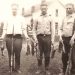 A vintage black and white photo shows three young men with rifles and game birds standing outside on a farm.