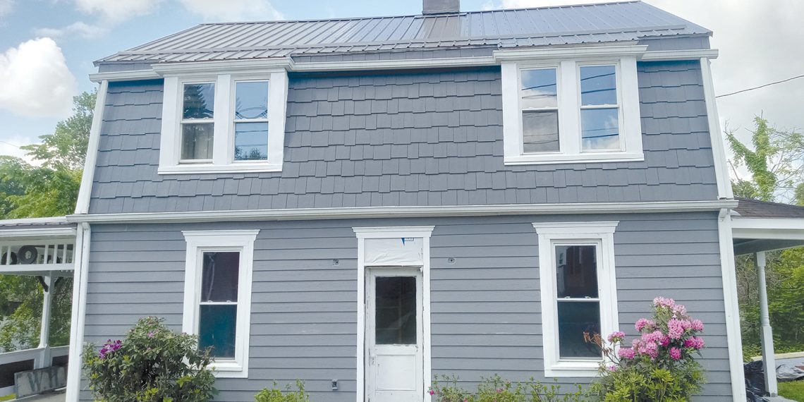 A gray farmhouse with white trim, a metal roof, and blooming bushes stands in a rural setting.