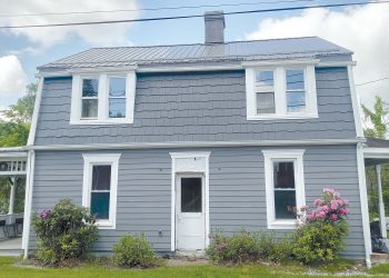 A gray farmhouse with white trim, a metal roof, and blooming bushes stands in a rural setting.