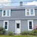 A gray farmhouse with white trim, a metal roof, and blooming bushes stands in a rural setting.