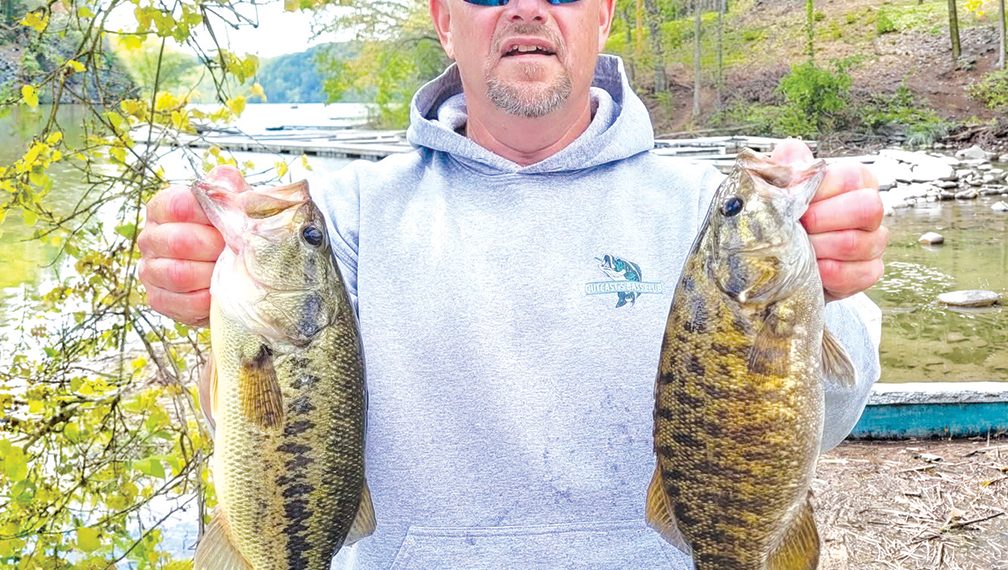 A man stands beside a lake holding two large bass he caught while fishing.
