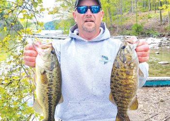 A man stands beside a lake holding two large bass he caught while fishing.