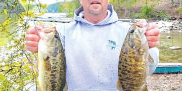 A man stands beside a lake holding two large bass he caught while fishing.