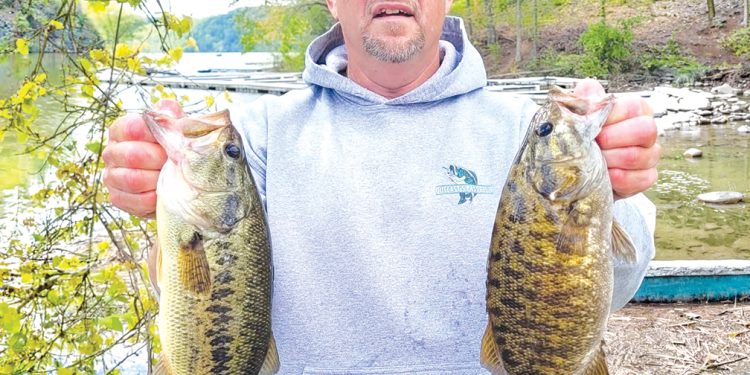 A man stands beside a lake holding two large bass he caught while fishing.