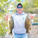 A man stands beside a lake holding two large bass he caught while fishing.