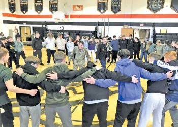 Students and JROTC cadets stand together in a circle during a group activity inside a school gymnasium.
