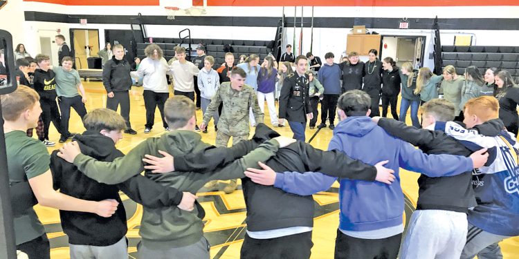 Students and JROTC cadets stand together in a circle during a group activity inside a school gymnasium.