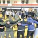 Students and JROTC cadets stand together in a circle during a group activity inside a school gymnasium.