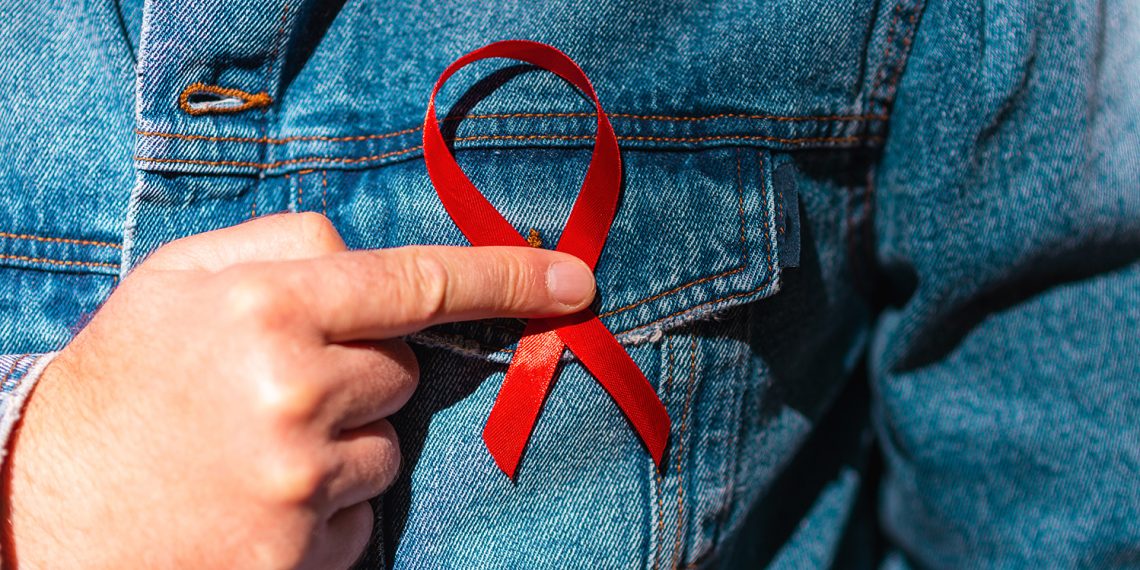 Man wearing Denim Jacket holding a red AIDS awareness ribbon on his chest.