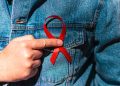 Man wearing Denim Jacket holding a red AIDS awareness ribbon on his chest.