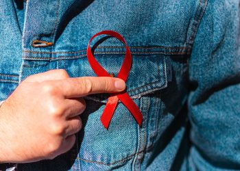 Man wearing Denim Jacket holding a red AIDS awareness ribbon on his chest.