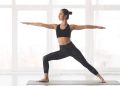 A woman performs a yoga pose on a mat inside a bright studio with large windows.