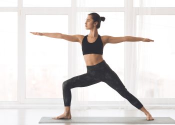 A woman performs a yoga pose on a mat inside a bright studio with large windows.