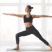 A woman performs a yoga pose on a mat inside a bright studio with large windows.