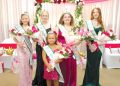 Five crowned fair royalty winners wearing sashes pose with bouquets in front of a pink and floral celebration backdrop.