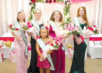 Five crowned fair royalty winners wearing sashes pose with bouquets in front of a pink and floral celebration backdrop.