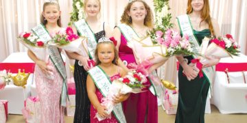 Five crowned fair royalty winners wearing sashes pose with bouquets in front of a pink and floral celebration backdrop.