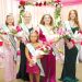 Five crowned fair royalty winners wearing sashes pose with bouquets in front of a pink and floral celebration backdrop.