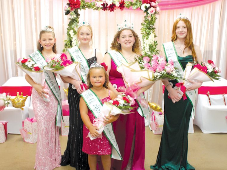 Five crowned fair royalty winners wearing sashes pose with bouquets in front of a pink and floral celebration backdrop.