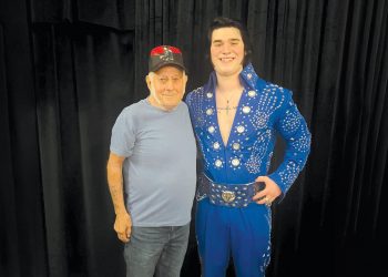 An older man stands beside an Elvis tribute artist in a blue rhinestone jumpsuit as they pose for a photo in front of a black curtain.
