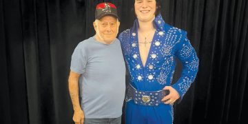 An older man stands beside an Elvis tribute artist in a blue rhinestone jumpsuit as they pose for a photo in front of a black curtain.
