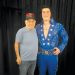 An older man stands beside an Elvis tribute artist in a blue rhinestone jumpsuit as they pose for a photo in front of a black curtain.