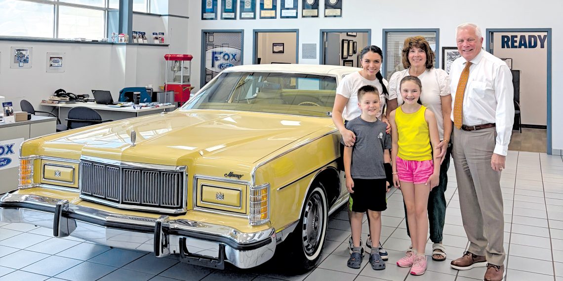 A family stands beside a vintage yellow Mercury convertible inside a Ford dealership showroom.