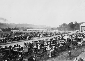 A historic black and white photo shows the Greene County Fairgrounds crowded with early automobiles, horse drawn buggies, spectators, and a grandstand beside the track.