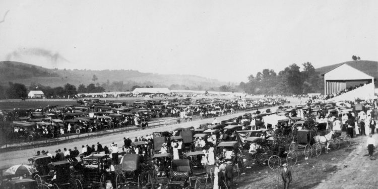 A historic black and white photo shows the Greene County Fairgrounds crowded with early automobiles, horse drawn buggies, spectators, and a grandstand beside the track.