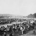 A historic black and white photo shows the Greene County Fairgrounds crowded with early automobiles, horse drawn buggies, spectators, and a grandstand beside the track.