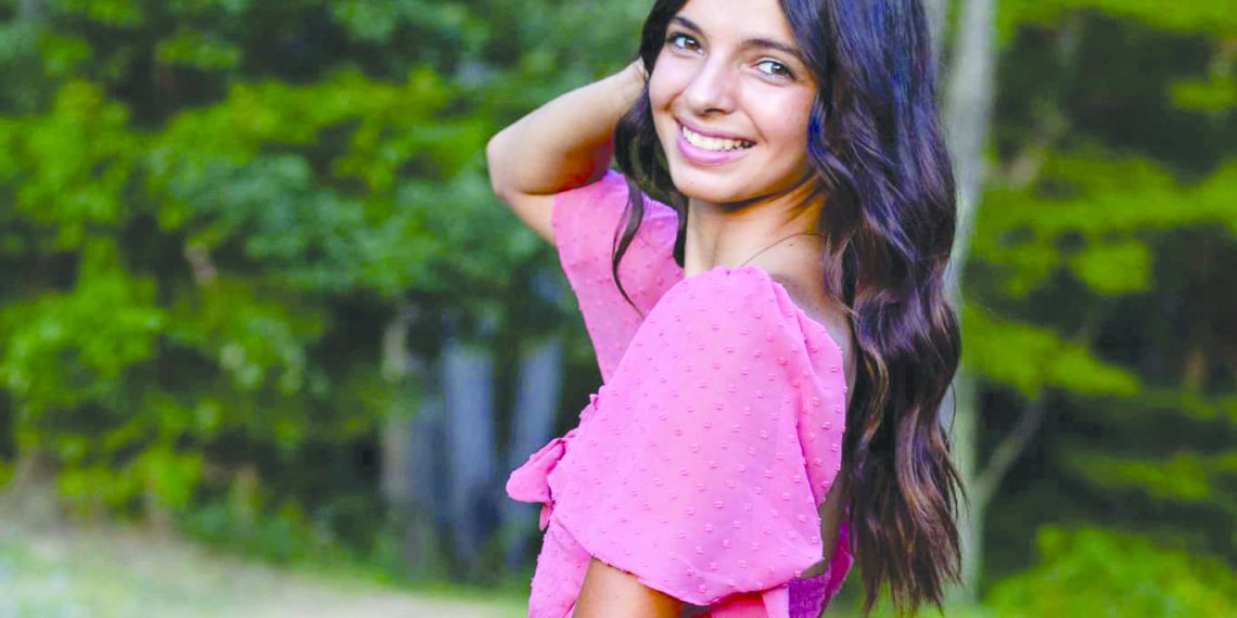 A smiling young woman in a pink dress poses beside a pond with trees in the background.