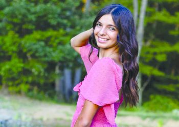 A smiling young woman in a pink dress poses beside a pond with trees in the background.