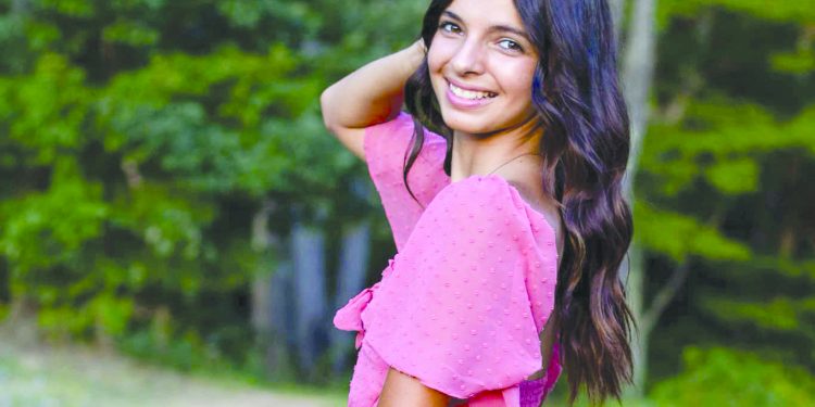 A smiling young woman in a pink dress poses beside a pond with trees in the background.