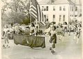 A vintage photo shows young parade participants carrying an American flag and a banner as they march down a town street lined with spectators and classic cars.