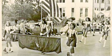 A vintage photo shows young parade participants carrying an American flag and a banner as they march down a town street lined with spectators and classic cars.
