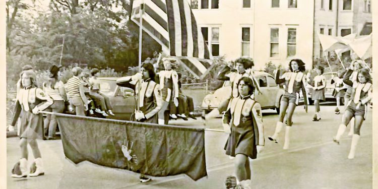 A vintage photo shows young parade participants carrying an American flag and a banner as they march down a town street lined with spectators and classic cars.