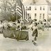 A vintage photo shows young parade participants carrying an American flag and a banner as they march down a town street lined with spectators and classic cars.