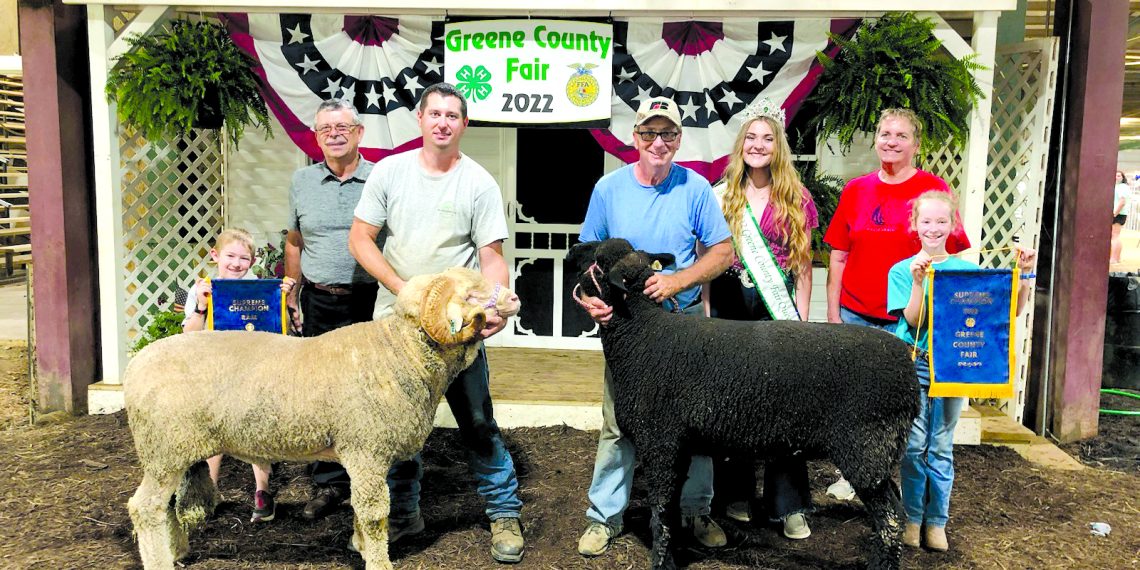 Exhibitors, family members, and fair royalty pose with a champion ram and champion sheep while children display championship banners at the Greene County Fair.