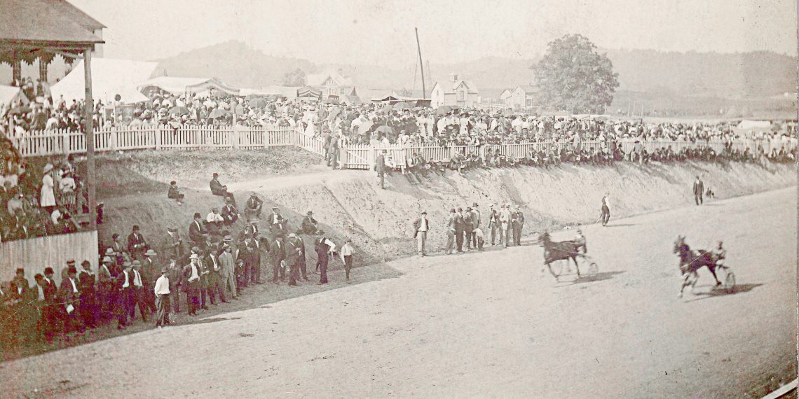 A historic photograph shows spectators watching a harness horse race on a dirt track at an early county fair.