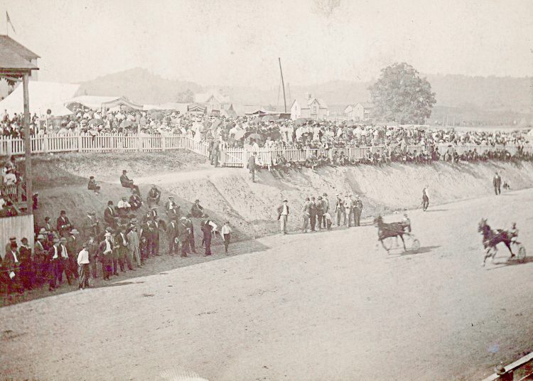 A historic photograph shows spectators watching a harness horse race on a dirt track at an early county fair.