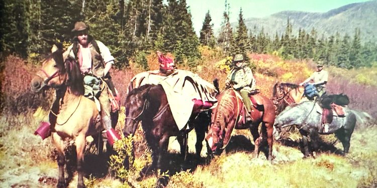 A group of hunters on horseback gathers in a mountain meadow with pack horses carrying harvested game during a backcountry hunt.