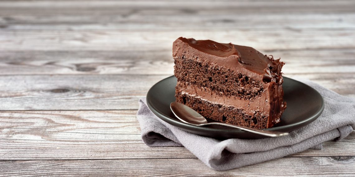A slice of chocolate layer cake with chocolate frosting sits on a black plate with a spoon on a rustic wooden table.