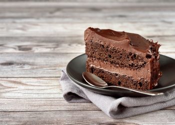 A slice of chocolate layer cake with chocolate frosting sits on a black plate with a spoon on a rustic wooden table.