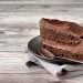 A slice of chocolate layer cake with chocolate frosting sits on a black plate with a spoon on a rustic wooden table.