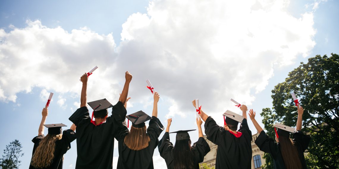 Rear portrait of group students raise fists hold diploma mortarboard gown graduate college building outdoors.
