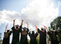 Rear portrait of group students raise fists hold diploma mortarboard gown graduate college building outdoors.