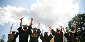 Rear portrait of group students raise fists hold diploma mortarboard gown graduate college building outdoors.