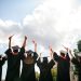 Rear portrait of group students raise fists hold diploma mortarboard gown graduate college building outdoors.