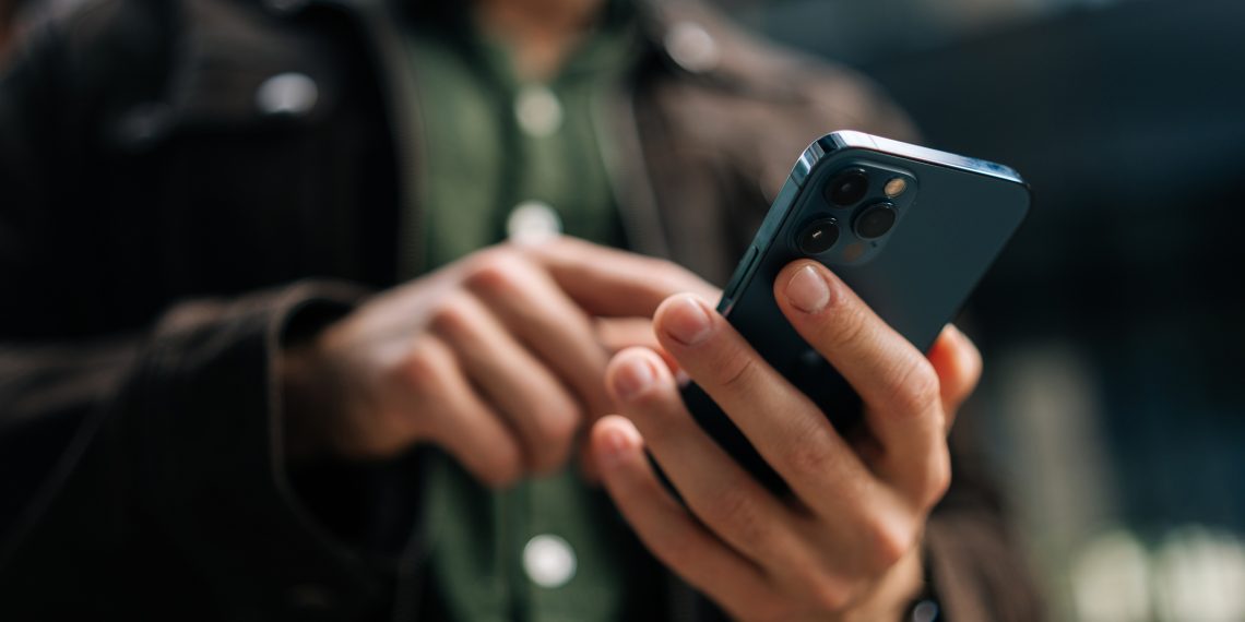 Close-up hands of unrecognizable man holding and using smartphone standing on city street, browsing internet, checking social media, using mobile application. Concept of modern communication.